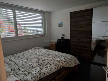 Cette chambre confortable dispose d'une grande fenêtre avec des stores, d'un lit avec une couette à motifs, d'une armoire sombre avec des miroirs, et d'un décor minimaliste avec des murs clairs.