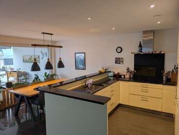 Cette image montre une cuisine ouverte moderne et une salle à manger avec des armoires jaunes, des comptoirs noirs, des lampes suspendues, des plantes, des œuvres d'art et une vue sur un balcon.