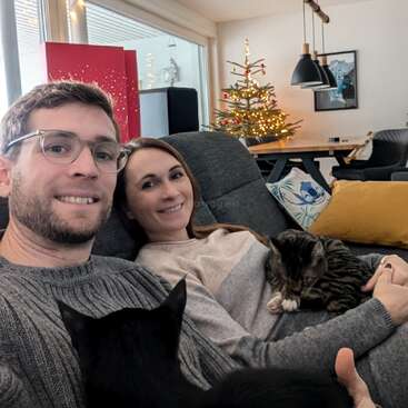 Un couple souriant est assis sur un canapé confortable avec deux chats. Derrière eux se trouvent un arbre de Noël décoré, des lumières festives et un salon moderne et accueillant.