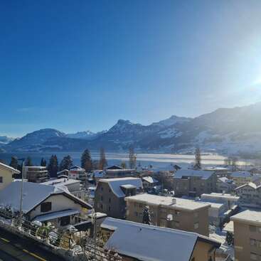 A serene winter scene of a snowy village beneath clear blue skies. Majestic mountains and a bright sun overlook the peaceful houses and shimmering frozen lake.
