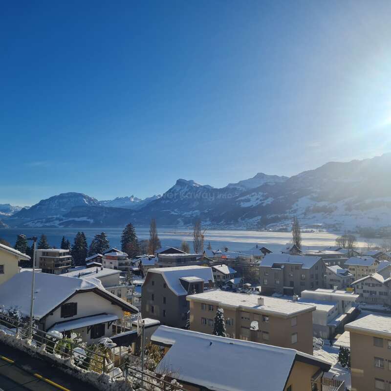 Scène hivernale sereine d'un village enneigé sous un ciel bleu clair. Des montagnes majestueuses et un soleil radieux surplombent les maisons paisibles et le lac gelé scintillant.
