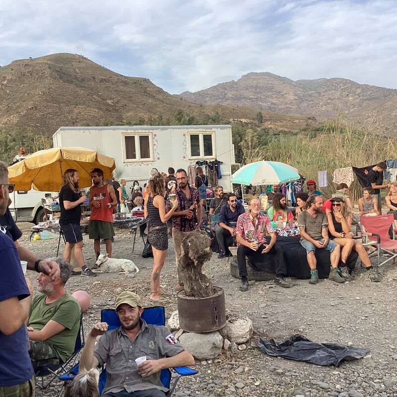 L'image représente un rassemblement de personnes dans un cadre désertique, avec une caravane blanche et des montagnes en arrière-plan, dans une atmosphère détendue et décontractée.