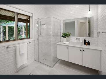 Esse banheiro moderno tem azulejos brancos, chuveiro com box de vidro, espelho grande, penteadeira elegante, pia com vaso, planta de interior, toalheiro e muita luz natural.