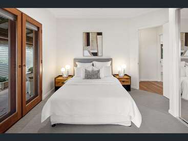 Um quarto moderno e minimalista com uma cama bem arrumada, mesas laterais de madeira, arte abstrata na parede, armário espelhado e portas duplas de vidro que levam a uma área externa.