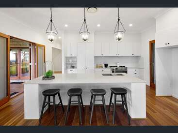 Essa cozinha moderna apresenta uma grande ilha branca, quatro banquetas pretas, luzes pendentes elegantes, pisos de madeira, armários minimalistas, aparelhos de aço inoxidável e muita luz natural.