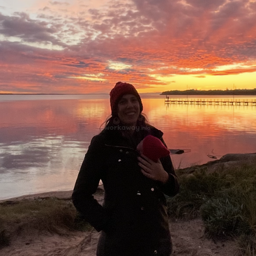 Uma mulher está perto de um lago calmo ao pôr do sol, usando um casaco escuro e um chapéu vermelho. Nuvens vibrantes em laranja e rosa preenchem o dramático céu noturno.