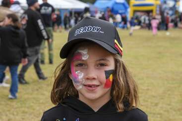 A smiling child wearing a "DEADLY" hat stands outdoors at an event, with colorful face paint and people milling in the background on a grassy field.