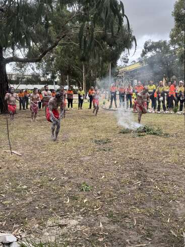 A group of Indigenous dancers, painted and wearing red cloth, perform a traditional dance, while a crowd of onlookers in work uniforms watch respectfully outdoors.
