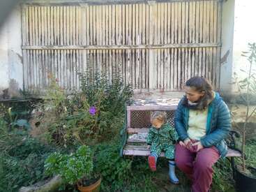 Uma mulher e uma criança sentam-se em um banco velho em um jardim com plantas crescidas. A parede atrás deles está desgastada e é de madeira.