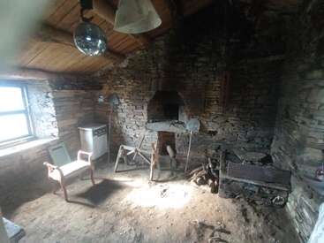 Esta imagem mostra uma sala de pedra antiga e rústica com um forno a lenha, móveis simples de madeira, uma bola de discoteca, piso de terra batida e uma janela que deixa entrar luz.