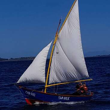 Un petit voilier bleu nommé "Swa Kalk" navigue sur une eau bleu foncé sous un ciel clair. Deux personnes portant des gilets de sauvetage naviguent ensemble sur le bateau.
