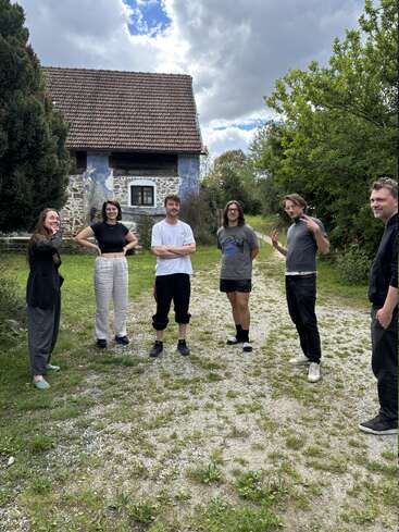 Six personnes se tiennent dehors sur un chemin de gravier, souriantes et prenant la pose. Derrière elles se dresse une maison rustique en pierre au toit de tuiles, entourée de verdure.