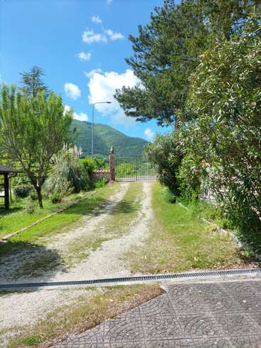 Un camino de grava se extiende a través de un exuberante jardín verde, bordeado de árboles y arbustos, que conduce a una entrada cerrada. Detrás, cielo azul y montañas.