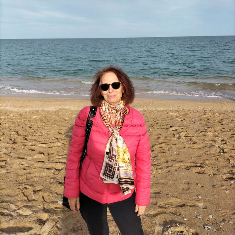 Una mujer en una playa de arena con una chaqueta rosa, un pañuelo estampado y gafas de sol. El mar y el cielo ofrecen un fondo tranquilo y pintoresco.