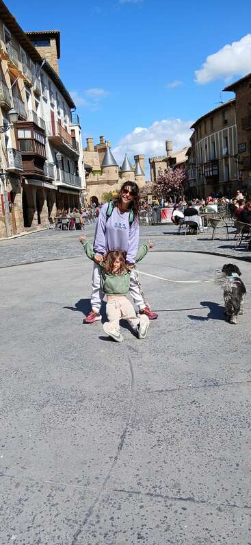 A woman and a child play joyfully in a picturesque town square, accompanied by a small dog. People sit at outdoor cafes, with castle towers behind.