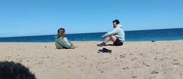 Two people sit on a sandy beach facing each other, wearing hoodies. The blue ocean and sky create a calm, peaceful atmosphere. Shoes lie nearby.