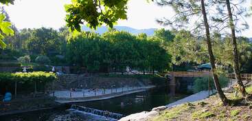 Uma cena tranquila à beira do rio, com árvores verdes e exuberantes, pessoas relaxando à beira da água, uma área de praia arenosa e uma ponte de madeira. Dá pra ver as montanhas ao fundo.