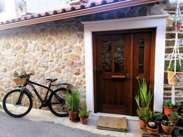 Uma casa rústica de pedra tem uma porta de madeira, cercada por vasos de plantas. Uma bicicleta com uma cestinha está encostada na parede, criando um clima aconchegante.