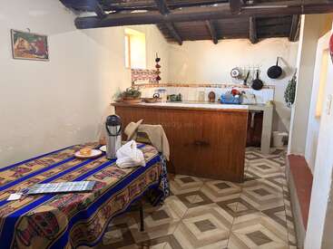 A cozy rustic kitchen features a wooden counter, patterned floor, colorful tablecloth, basic utensils, simple decor, open shelves, and a warm, inviting atmosphere with natural light.
