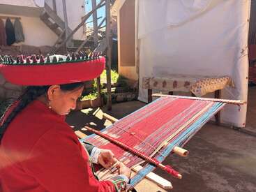 A woman in traditional Andean clothing weaves colorful threads using a backstrap loom outdoors, with sunlight illuminating her intricate, skilled handiwork in a peaceful setting.