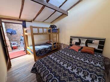 This cozy bedroom features a double bed, a wooden bunk bed, patterned bedding, and an open door revealing an outdoor space with a person and stone walls.