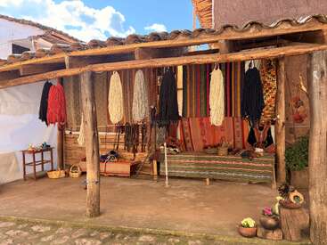 A rustic outdoor workspace displays colorful yarn hanging from a wooden beam, woven textiles, baskets, a bench, and vibrant artisan decorations beneath a traditional tiled roof.