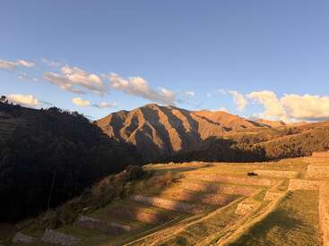 Golden sunlight illuminates ancient terraced fields, nestled against green hills and dramatic, shadowed mountains under a clear sky with scattered white clouds in the background.