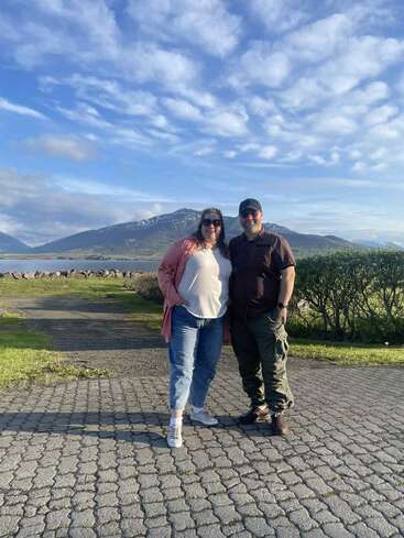 Un couple se tient ensemble en plein air par une journée ensoleillée, entouré de belles montagnes, d'un lac et de verdure, avec un ciel bleu et des nuages au-dessus d'eux.