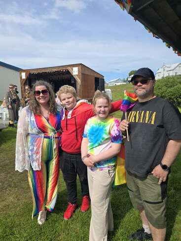 Une famille souriante pose en plein air lors d'une manifestation de la Fierté, vêtue de tenues arc-en-ciel et tie-dye. Le temps est ensoleillé et les gens célèbrent ensemble l'inclusion.
