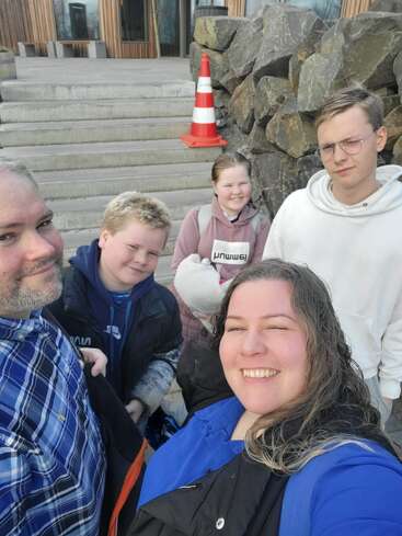 L'image montre une famille de cinq personnes posant pour un selfie sur des marches en béton, avec un cône de signalisation derrière eux et un mur de pierre à droite.