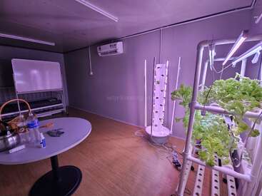 Esta imagen muestra una granja hidropónica de interior con verduras de hoja verde bajo luces LED. Hay una mesa con artículos, una pizarra, aire acondicionado y suelo de madera.