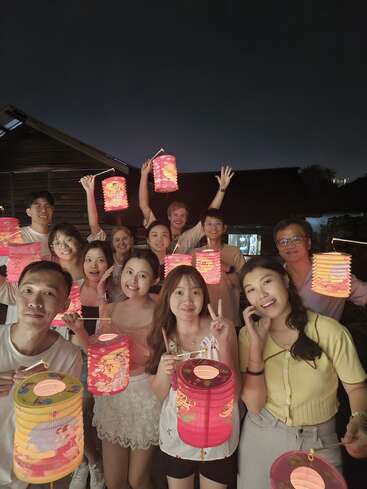 Un grupo de personas felices posan juntas por la noche, sosteniendo farolillos de colores brillantes, sonriendo y celebrando al aire libre bajo el cielo oscuro, probablemente durante un evento festivo.