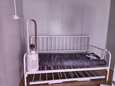 Una habitación minimalista presenta un somier de metal blanco con un colchón oscuro. Sobre la cama hay cables, y a su lado hay un purificador de aire sin aspas.