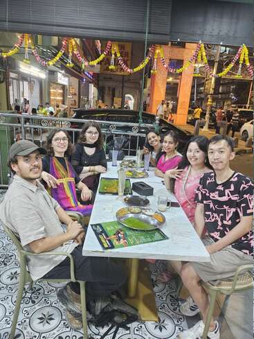 Sept amis sont assis autour d'une table de restaurant, dégustant des plats et des boissons. Des guirlandes festives sont suspendues au-dessus d'eux. L'arrière-plan montre une rue animée de nuit.