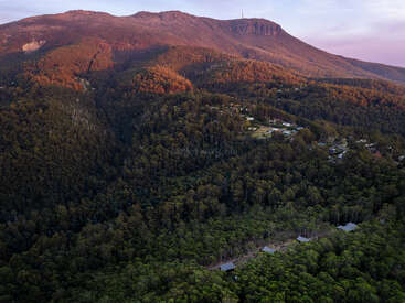 Vista aérea de uma paisagem montanhosa coberta de mata exuberante ao pôr do sol, com casas espalhadas entre as árvores e um pico rochoso bem visível ao longe.