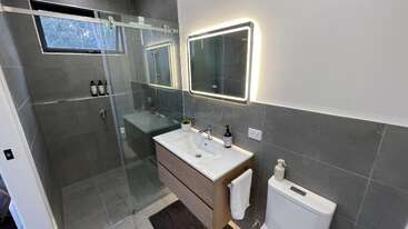 Esta casa de banho moderna tem um box de vidro, paredes revestidas com azulejos cinza, um lavatório suspenso com duas pias, espelho iluminado, vaso sanitário e uma decoração minimalista com produtos de higiene pessoal à vista.
