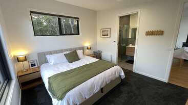 Este quarto aconchegante tem uma cama grande com detalhes em verde, mesinhas de cabeceira e abajures, uma janela com vista para a vegetação lá fora, piso alcatifado e um banheiro moderno ao lado.