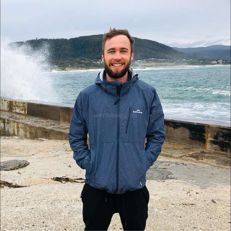 Um homem sorridente, de barba, está à beira-mar, com as mãos nos bolsos. As ondas quebram contra uma barreira de concreto. Ao fundo, veem-se colinas e um céu nublado.