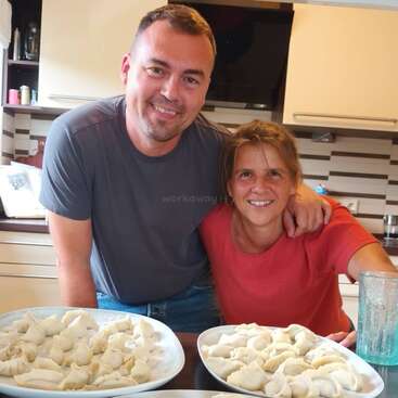 The image shows a man and woman posing behind a kitchen counter with two plates of dumplings, smiling at the camera.