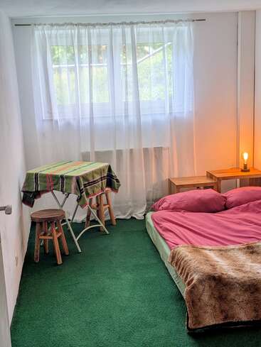 Esta acogedora habitación cuenta con moqueta verde, una cama baja con ropa de cama rosa, una manta de peluche, una mesita con taburetes, cortinas transparentes y una lámpara que crea un ambiente cálido.