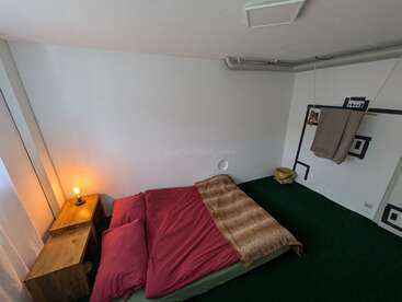 Un acogedor dormitorio de estilo minimalista cuenta con una cama doble con ropa de cama roja, una mesita de noche de madera, una lámpara encendida, una alfombra verde, un cuadro en la pared, una toalla colgada y una decoración sencilla.