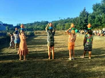 Six people, some blindfolded, balance clay pots on their heads in an open field while a seated audience watches, under a clear blue sky.