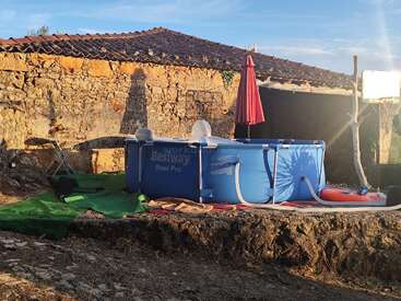 A blue above-ground pool stands on green artificial grass beside an old stone building, with a red umbrella, chair, and rustic, sunlit surroundings.