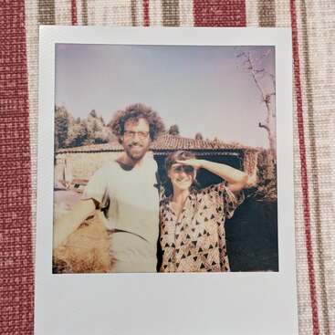 Una foto Polaroid muestra a dos personas sonrientes al aire libre, una con gafas y la otra con gafas de sol, de pie juntas sobre un fondo rústico con un tejido a rayas.