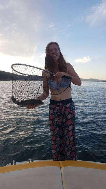 Uma mulher está em um barco, segurando uma rede de pesca com um peixe dentro, em um corpo de água sereno e um céu azul com nuvens.