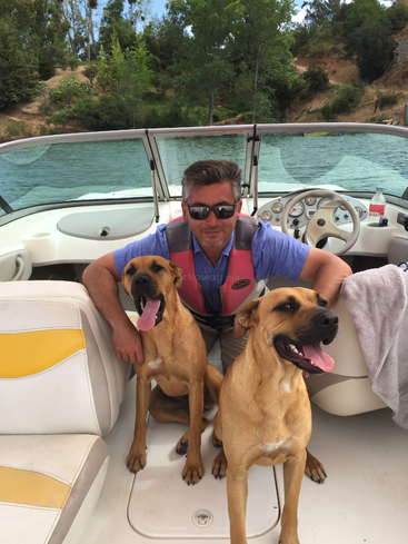 A imagem mostra um homem sentado em um barco com dois cães marrons em um lago, usando um colete salva-vidas e cercado por árvores.