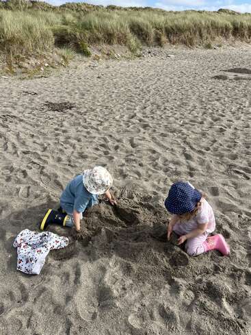 Dos niños pequeños con sombrero y botas están sentados en una playa de arena, cavando en la arena. Las dunas de hierba y el cielo azul aparecen al fondo.