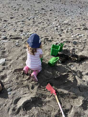 Un niño con sombrero azul y botas rosas juega en una playa de arena con regaderas verdes y una pala roja, disfrutando de un día soleado.
