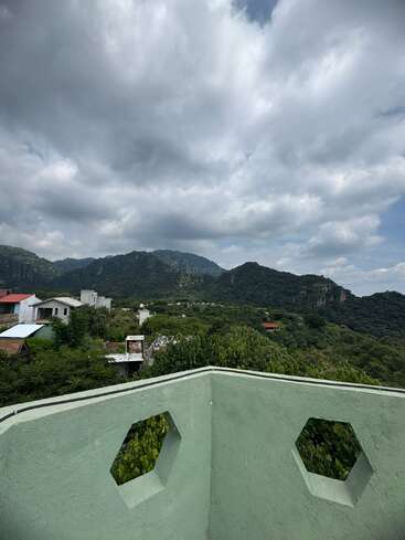 Una serena vista de montaña bajo un cielo nublado, un exuberante paisaje verde, casas dispersas y un muro verde claro con ventanas hexagonales en primer plano. Ambiente apacible.