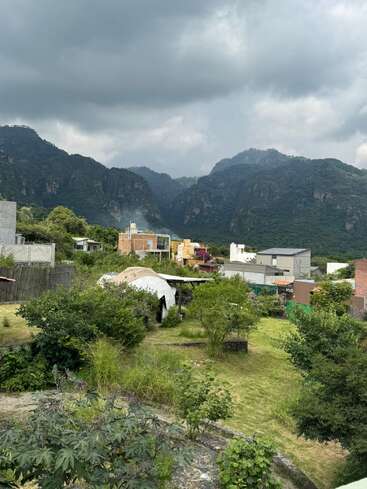Un pequeño barrio rodeado de exuberante vegetación, con montañas al fondo. Cielos oscuros y nublados se ciernen sobre el lugar, creando una atmósfera dramática sobre la tranquila escena.